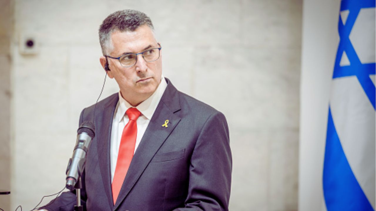 Na foto, está o Ministro das Relações Exteriores de Israel, Gideon Sa'ar, Homem de meia-idade, de pele clara e cabelos curtos grisalhos, está em pé diante de um microfone, vestindo terno escuro com camisa branca e gravata vermelha. Ele usa óculos e tem um ponto eletrônico na orelha esquerda. Na lapela do paletó, há um laço amarelo. O homem olha para o lado com expressão séria. Ao fundo, parcialmente visível à direita da imagem, está uma bandeira de Israel. O ambiente é interno, com parede de pedra clara ao fundo.