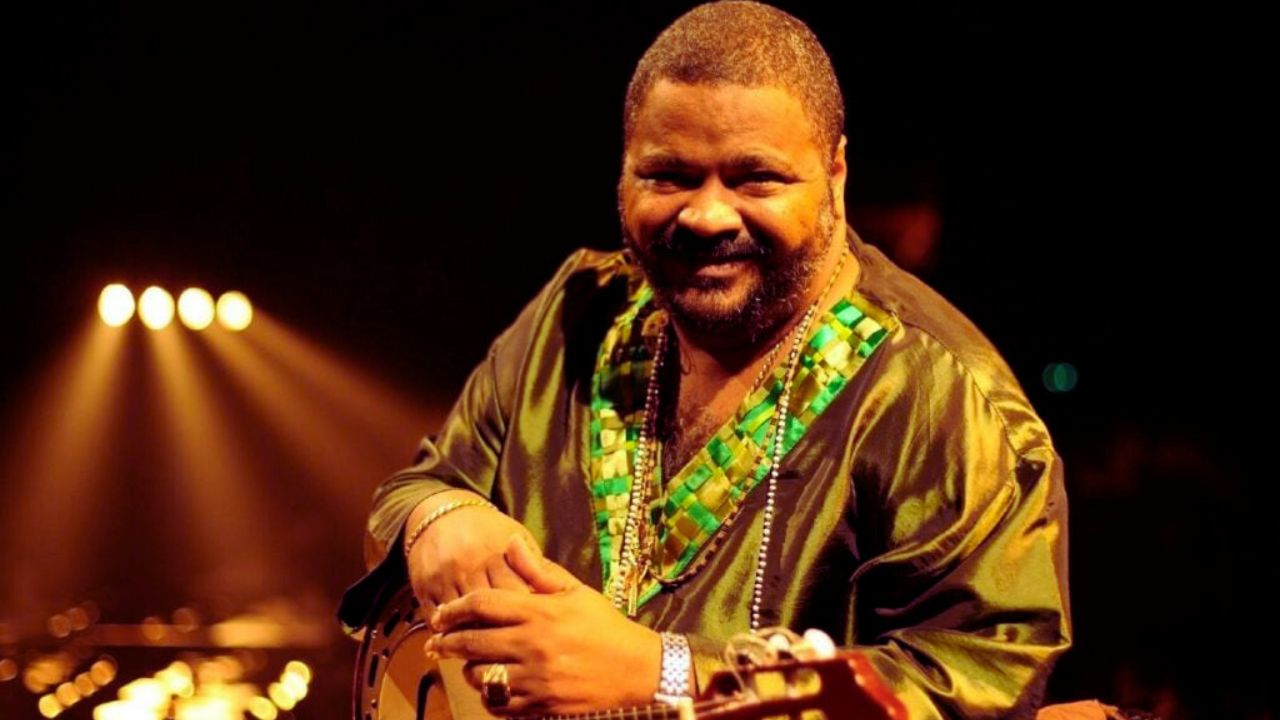 Na imagem, o sambista Arlindo Cruz está sentado e sorri levemente para a câmera. Ele veste uma túnica de seda verde com detalhes brilhantes em tons de verde e dourado na gola e nos punhos. Sua mão esquerda apoia um cavaquinho, parcialmente visível. Ao fundo, há luzes desfocadas e quentes, criando um clima acolhedor e intimista, típico de apresentações musicais em palco.